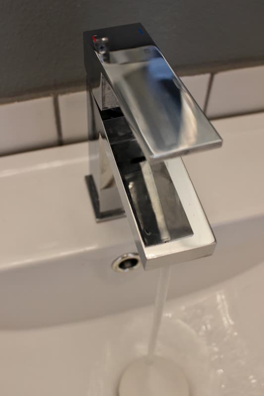 A modern waterfall faucet running on a bathroom sink against a white subway tile background.