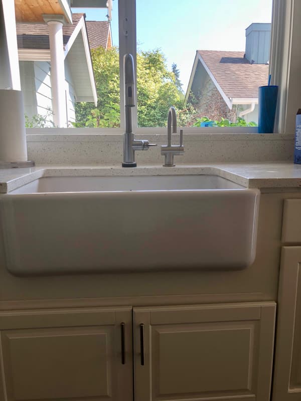 A farmhouse apron-front kitchen sink set in white cabinetry with a quartz countertop and a pull-down faucet, craftsman homes visible through the window.